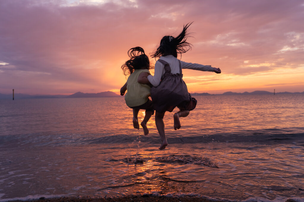 夕焼けの海を背景に、二人の娘がジャンプしている後ろ姿