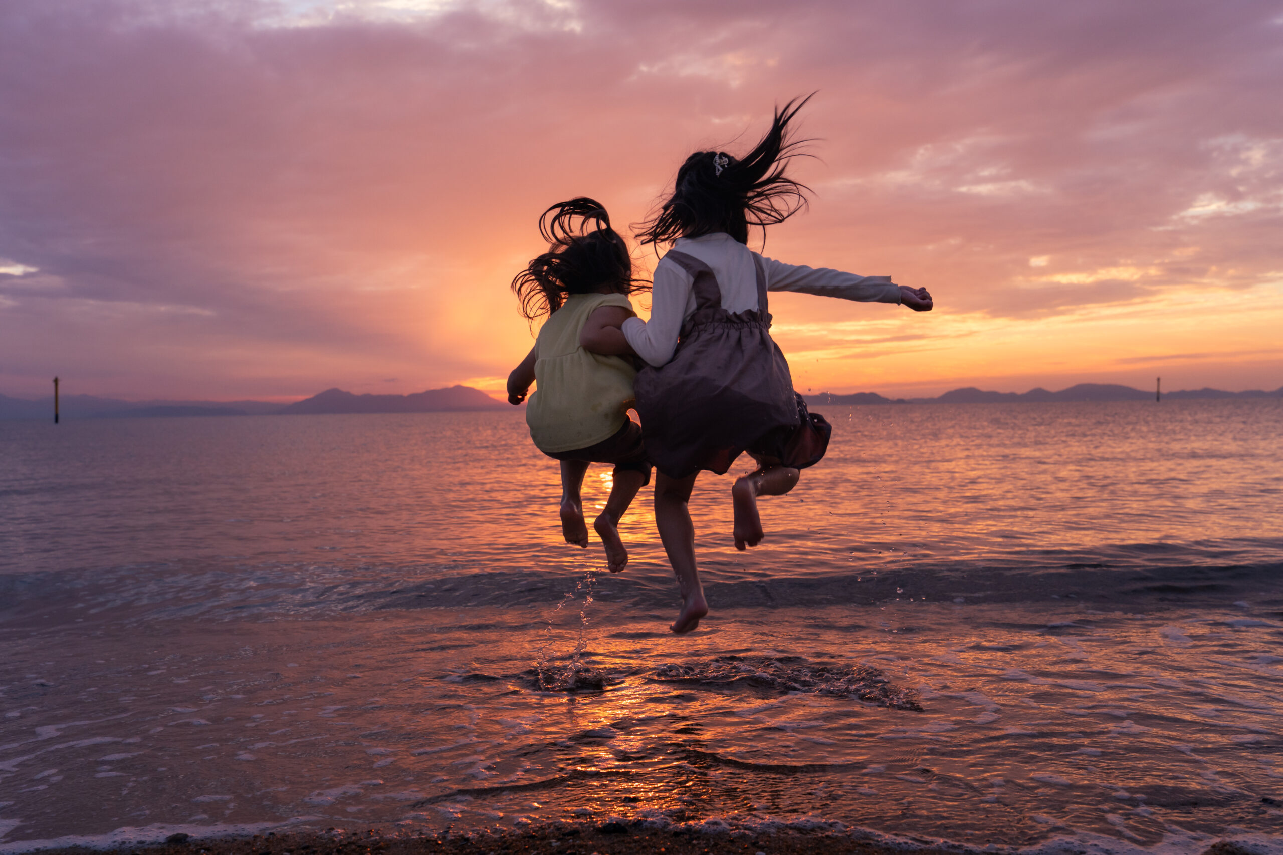 夕焼けの海を背景に、二人の娘がジャンプしている後ろ姿